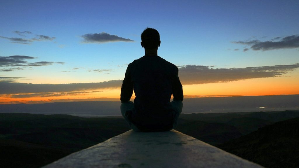 Person sitting on a ledge overlooking a distant scene with a sunset.
