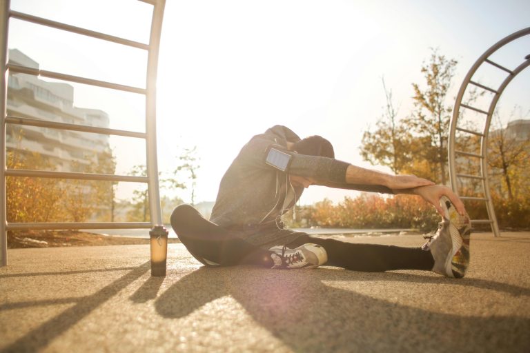 Guy stretching with one leg bent and reaching toward extended leg on the ground with the sun behind him.