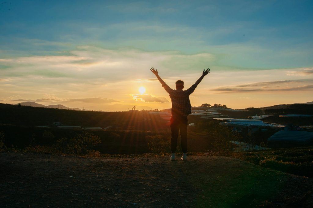 Person standing on hill with arms in the air facing the sun.