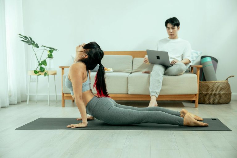 Woman doing yoga while a man is on a laptop on a couch.