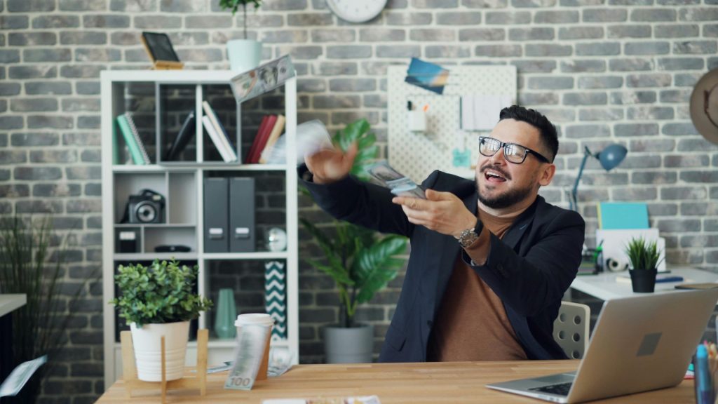 Man with glasses sitting at desk throwing around money with a smile.